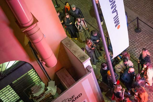 Visitors queue outside the House of Switzerland Milano during Milano Design Week, beside event signage and banners at the exhibition entrance in the evening.